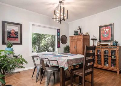 Homely dining room with a wooden table covered in a patterned tablecloth, surrounded by industrial metal chairs, rustic cabinet, and a modern chandelier.