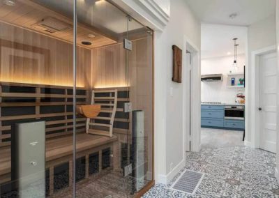 Modern home spa area with a wooden sauna, glass door, and a glimpse into a kitchen with patterned floor tiles and blue cabinetry.