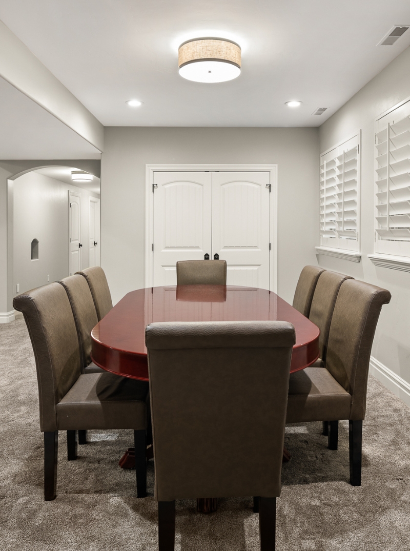 remodeled-interior-vertical-17 Spacious finished basement with plush carpeting, a large wooden coffee table, comfortable seating, and a dining set near windows with plantation shutters.