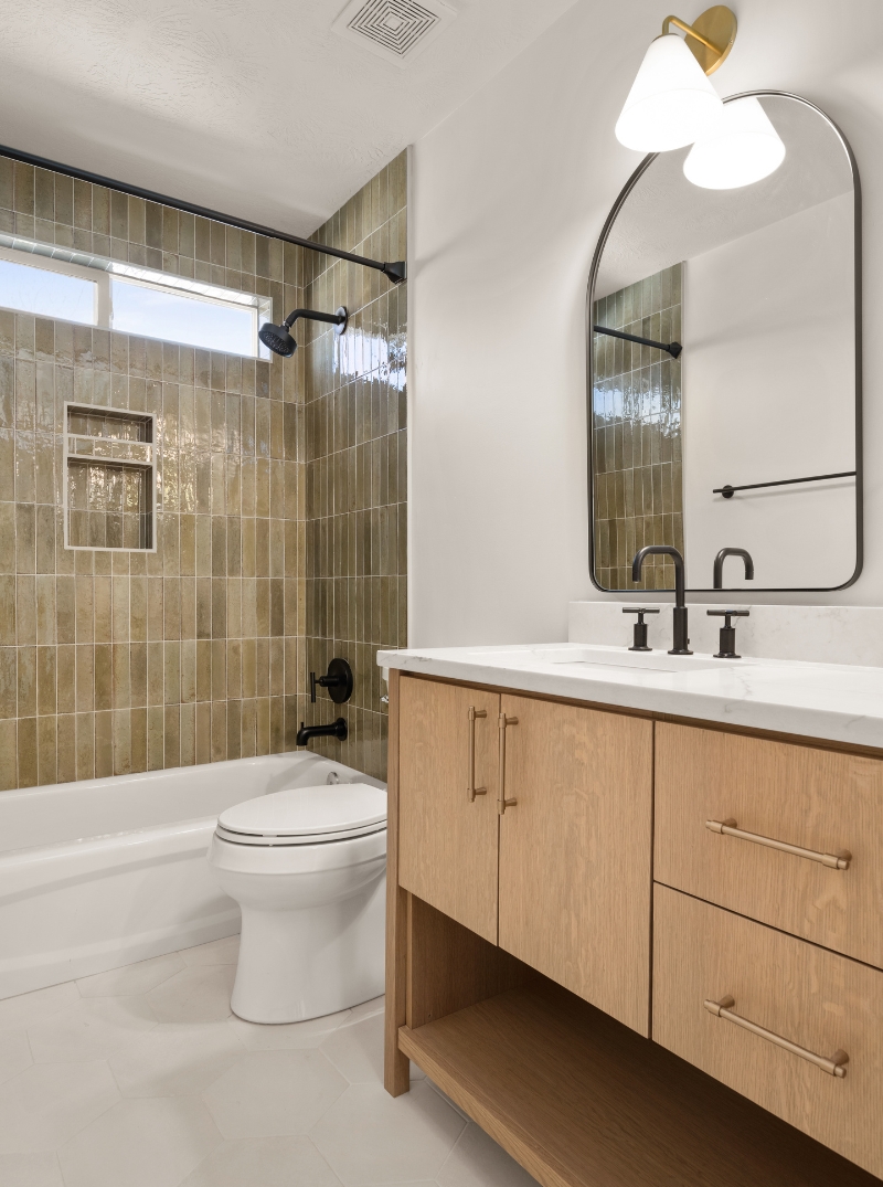 remodeled-interior-vertical-2 Modern bathroom interior with beige tiled walls, a white bathtub, wooden vanity with a white countertop, and a large mirror with black frame.