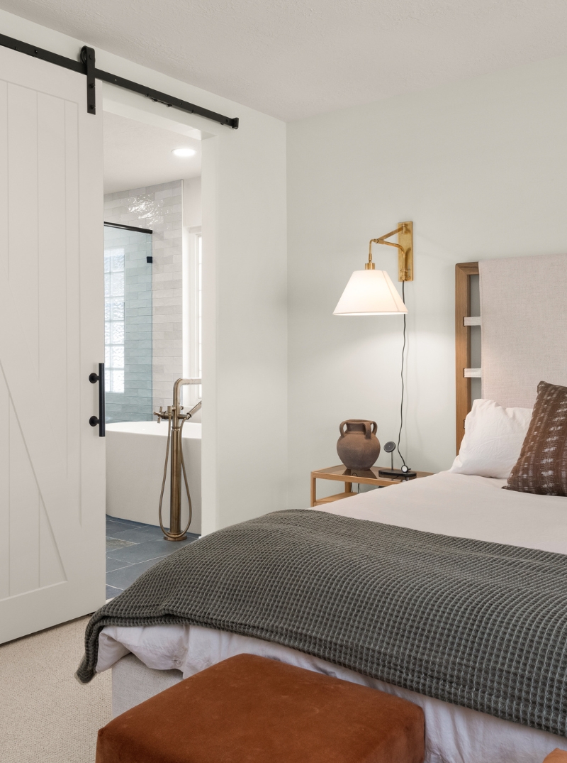 remodeled-interior-vertical-3 Elegant bedroom design featuring a barn door entrance to the bathroom, a mounted bedside lamp, and a comfortable bed with a grey knit blanket and a rust-colored ottoman at the foot.