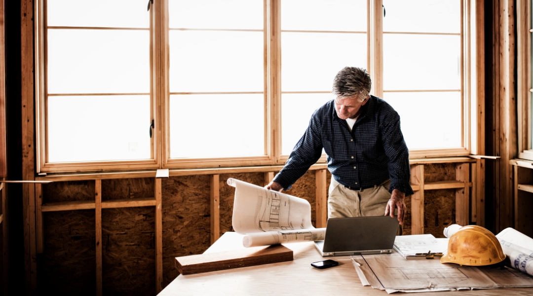 Experienced construction manager reviewing blueprints on a worktable at a new home construction site with large wooden framed windows in the background