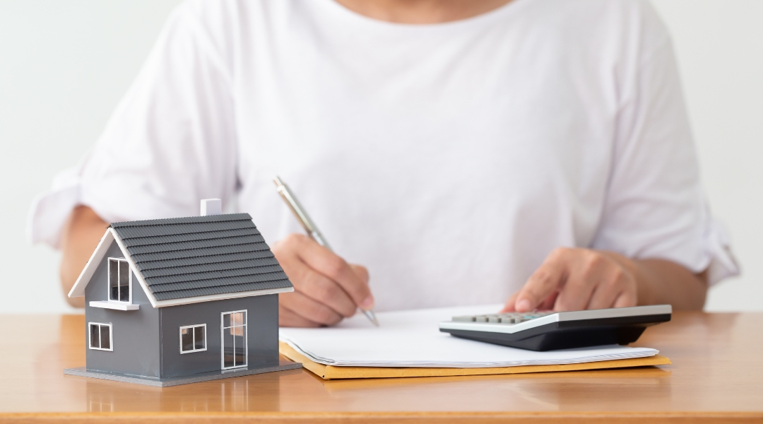 Homeowner planning finances for custom home building with a calculator, pen, and miniature house model on a desk